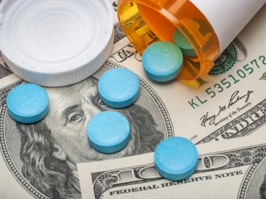 Blue pills spilling out of a prescription bottle on USD 100 dollar bills, photo by michaelquirk/Getty Images