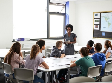 Female high school teacher standing by table with students teaching lesson, photo by Adobe Stock