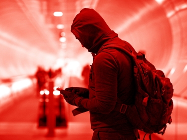A man in a subway tunnel wearing gloves and a hoodie, looking at a mobile phone. Photo by Lorado / Getty Images