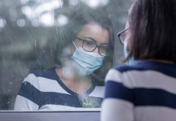 A woman wearing a face mask looks out a window in the rain