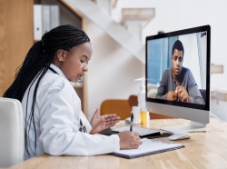 Black woman doctor writing notes during a video call with a Black male patient on a computer, photo by PeopleImages/Getty Images