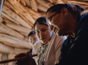 A senior Native American woman demonstrates weaving to two young women, photo by Hoptocopter / Getty Images