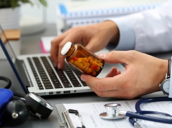 Close up of doctor holding a bottle of prescription pills in one hand and typing on a laptop with the other hand