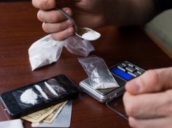 Close up of two hands using a spoon to put white powder in a small plastic bag on an electric scale, with money and phone on the table nearby