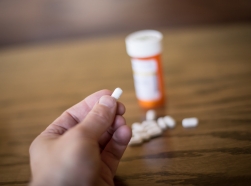 Hand holding a pill, with pills and a prescription bottle in the background, photo by stevehullphotography/Getty Images
