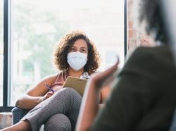 Seated female therapist wearing a mask, holding a clipboard, and facing her client, photo by SDI Productions/Getty Images