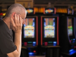 Depressed older white male gambler sitting in front of slot machines, photo by Ole/Adobe Stock