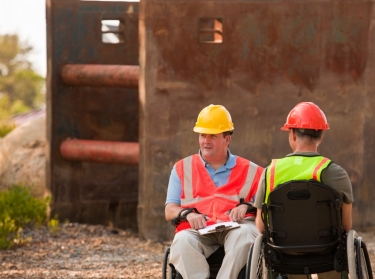 Two men onsite wearing hard hats in wheelchairs