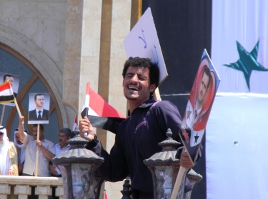 A man sits atop a lamp post waving pictures of Bashar Al Assad