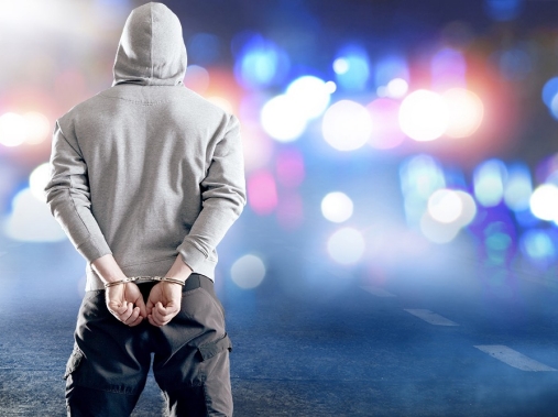 A young person in handcuffs faces police lights