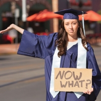 A female graduate in a cap and gown holding a Now What sign