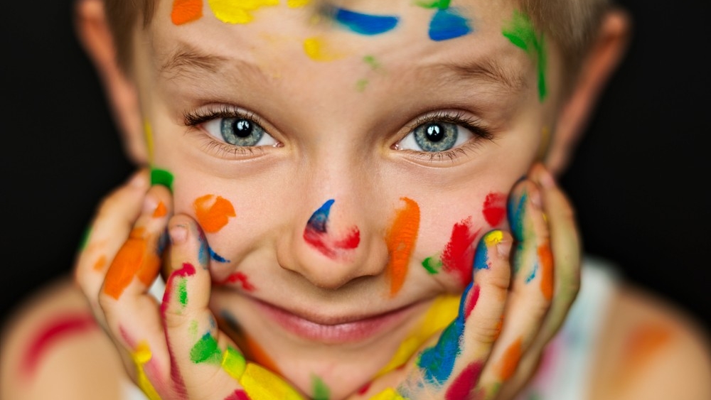 close-up of a child's face and hands, which are both covered in brightly-colored paint