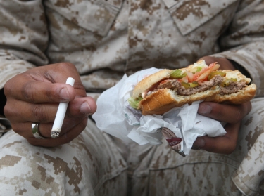 An individual in the fatigues worn by U.S. Marines holding a cigarette and hamburger