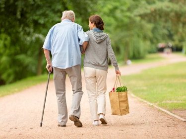 Female caregiver carrying groceries and walking along a dirt path with an elderly man