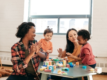 Two women sit at a table with two young children playing with blocks