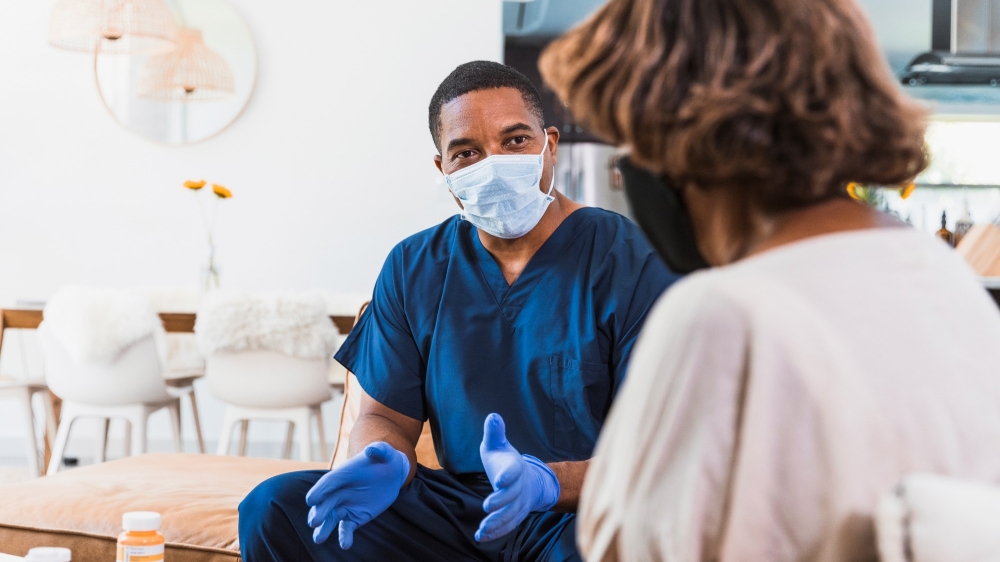 When the mid adult male home health nurse visits his senior adult female patient during the coronavirus outbreak, they both wear protective masks.  He also wears protective gloves.