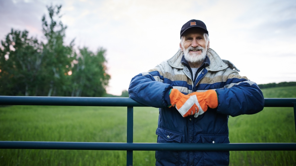 Older man standing at a farm fence and smiling at the camera. Photo by AJ_Watt/Getty Images