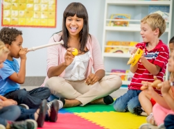 A group of preschool children sit with their teacher and play musical instruments