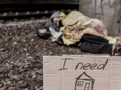 Homeless person sleeping on the ground, with a cardboard sign asking for a home in the foreground