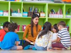Asian female teacher teaching diverse group of kids reading book sitting on library floor in classroom