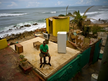 Roberto Figueroa Caballero sits on a small table in his home destroyed by Hurricane Maria, in La Perla neighborhood on the coast of San Juan, Puerto Rico, October 5, 2017, photo by Ramon Espinosa/AP Photos