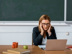 A stressed teacher sitting at her desk looking at a laptop, photo by Lightfield Studios/Adobe Stock