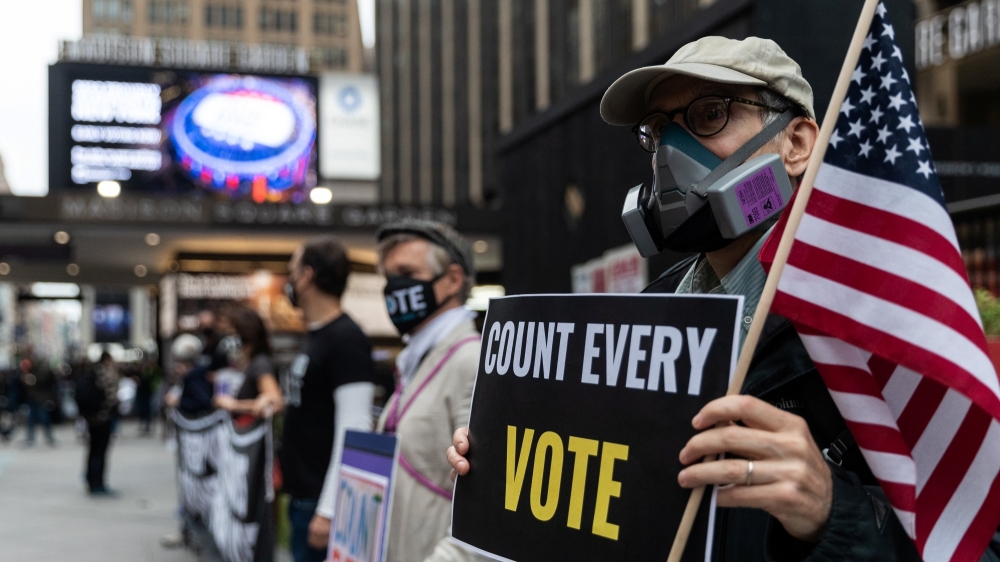 A man wearing a protective mask due to COVID-19 pandemic holds an American flag and a sign that says "count every vote"