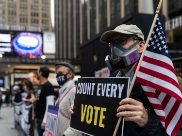 A man wearing a protective mask due to COVID-19 pandemic holds a sign outside Madison Square Garden, which is used as a polling station, on the first day of early voting in Manhattan, New York, October 24, 2020, photo by Jeenah Moon/Reuters