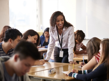 Female high school teacher standing by student table teaching lesson, photo by Monkey Business/AdobeStock