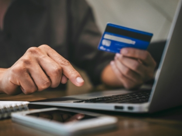 A person holds a credit card and types on a laptop while online shopping, photo by Ngampol/Adobe Stock
