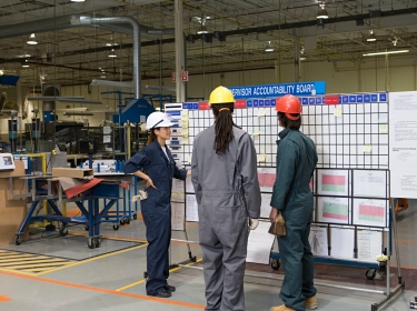 Three factory workers standing next to a message board in a factory