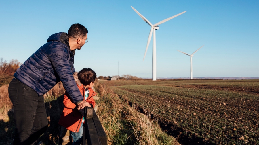 A man and young child leaning against a fence looking out into a field containing wind turbines. Photo by SolStock / Getty Images