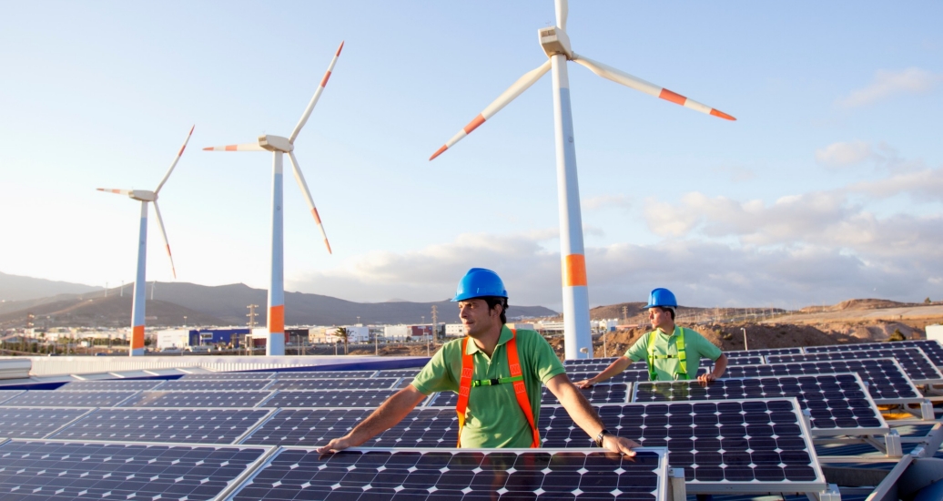 Two workers wearing hard hats on a solar farm with wind turbines in the background